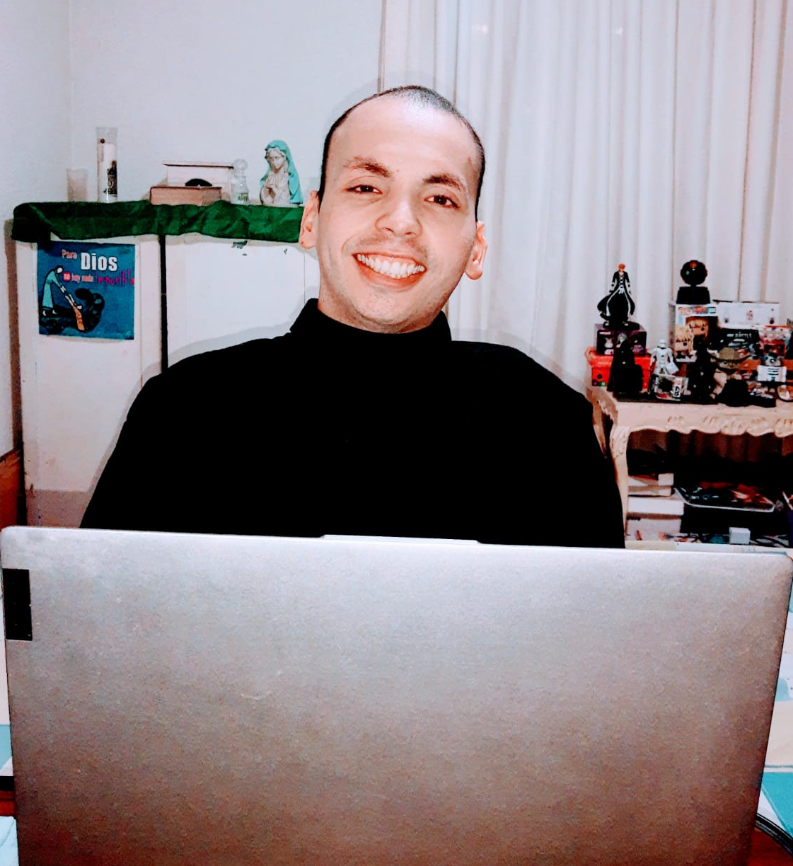 Imagen de Mikatholic sonriendo, tiene una camisa negra
        y enfrente de él su laptop gris, detras a su lado derecho tiene un letrero que dice Para Dios no hay nada imposible, una biblia,
        una estatua de la virgen María, las cenizas de su sobrino y una vela, detras a su lado izquierdo tiene muñecos de Star Wars, uno
        de Ichigo Kurosaki, un Funko pop de Minato y cartas tanto de Yu-gi-oh como de Pokemon