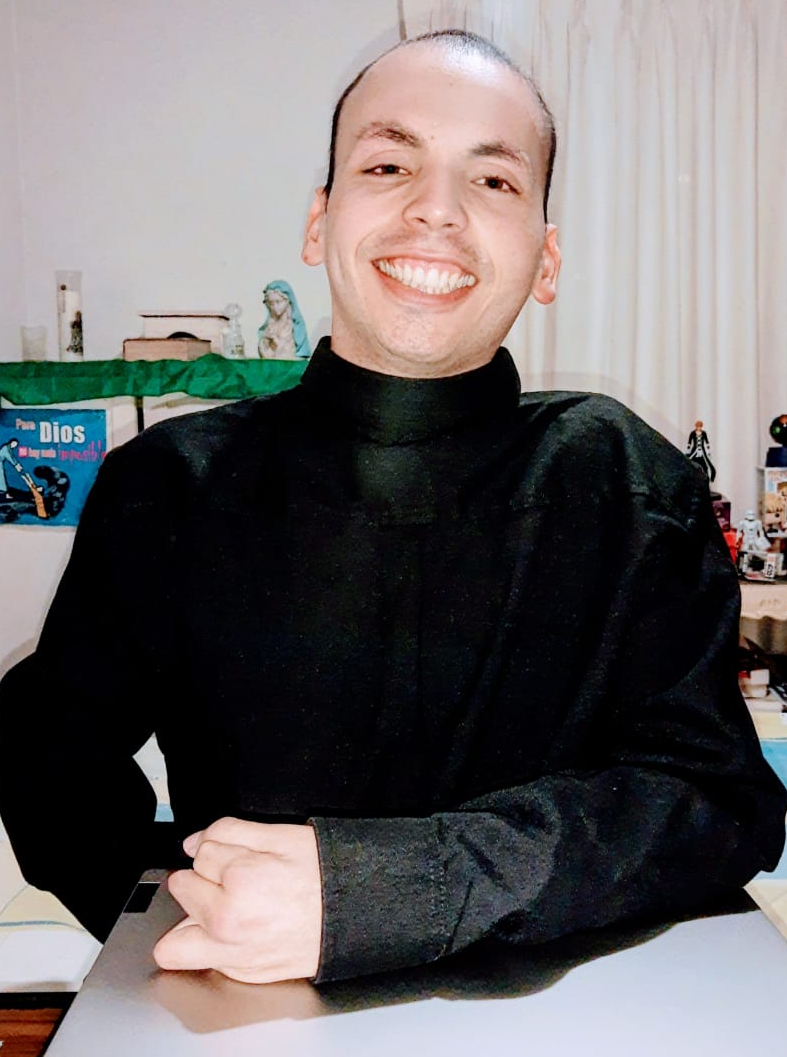 Imagen de Mikatholic sonriendo, tiene una camisa negra 
                y sobre su laptop cerrada tiene apoyando su brazo izquierdo, detras a su lado derecho tiene un letrero que dice Para Dios no hay nada imposible, una biblia, 
                una estatua de la virgen María, las cenizas de su sobrino y una vela, detras a su lado izquierdo se ve un muñeco de 
                Ichigo Kurosaki y un Funko pop de Minato
